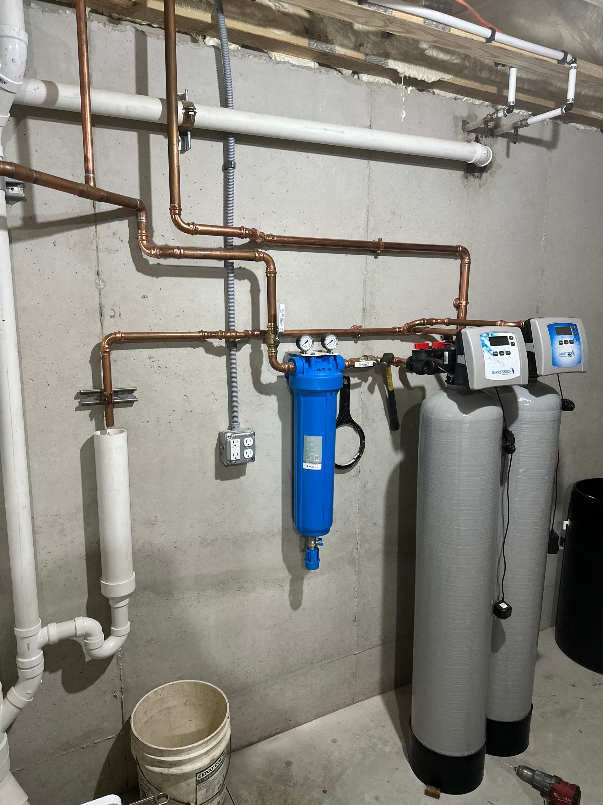 Basement utility wall with copper pipes, blue filter canister, and two water treatment tanks.