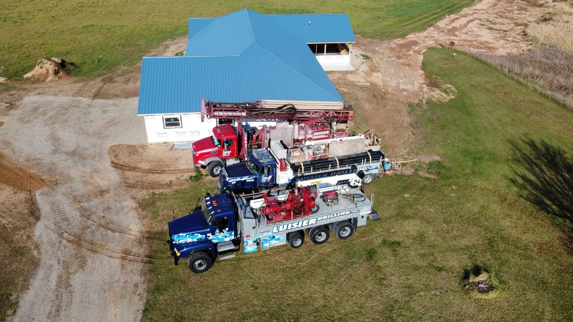Aerial view of two red and blue tractors with farm equipment beside a blue-roofed barn on a grassy field.