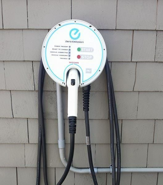 Electric vehicle charger mounted on a gray, wooden building. White and blue circular charging station with two cables.