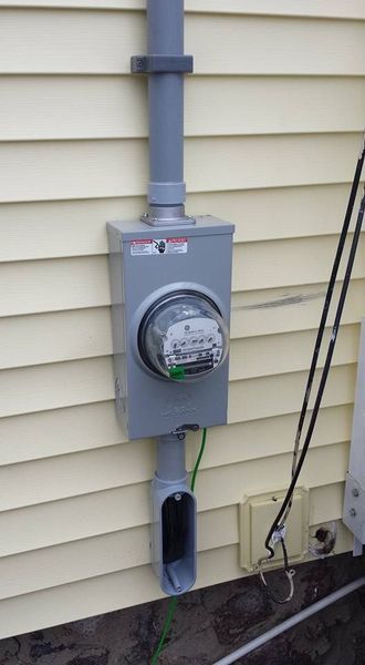 Electric meter box and conduit on a beige-sided house. Gray metal box with meter, vertical gray conduit, and exposed green ground wire.