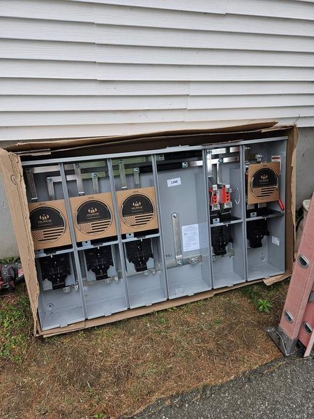 Electrical meter bank, gray metal, still in cardboard box, on the ground near a house.