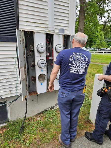 Man in blue uniform inspects a multi-meter electrical box on a building exterior. Another person in uniform is present.