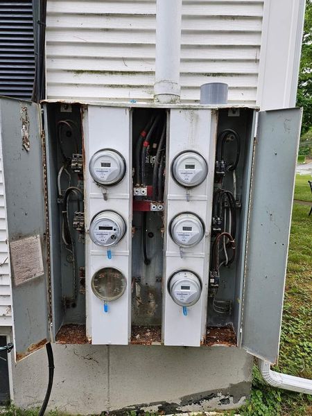 Gray metal electrical meter box on a white house, containing multiple meters.