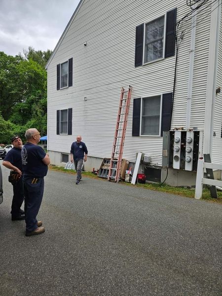 Three men by a white building with electrical boxes and a ladder. Two men stand, one walks towards them.