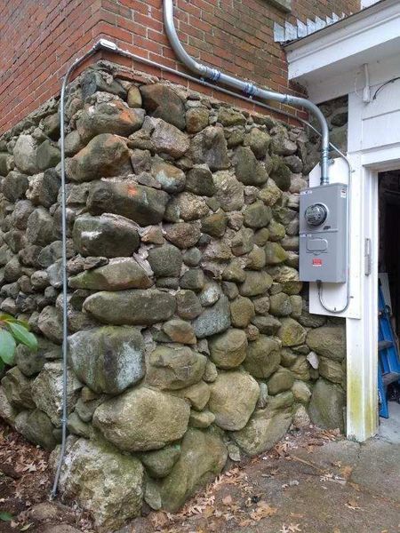 Electrical meter box mounted on a stone wall next to a white door frame. Conduit runs along the wall.