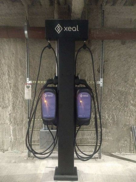 Black Xeal EV charging station with two chargers, mounted on a pedestal against a concrete wall.