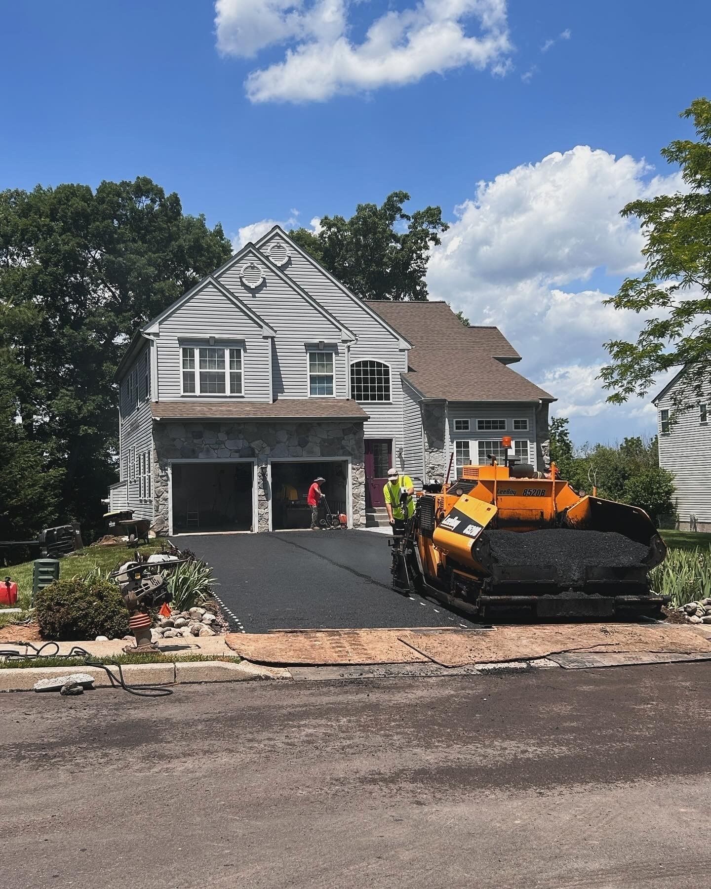 Workers operate a yellow paving machine to lay new asphalt on a residential driveway in front of a two-story house.