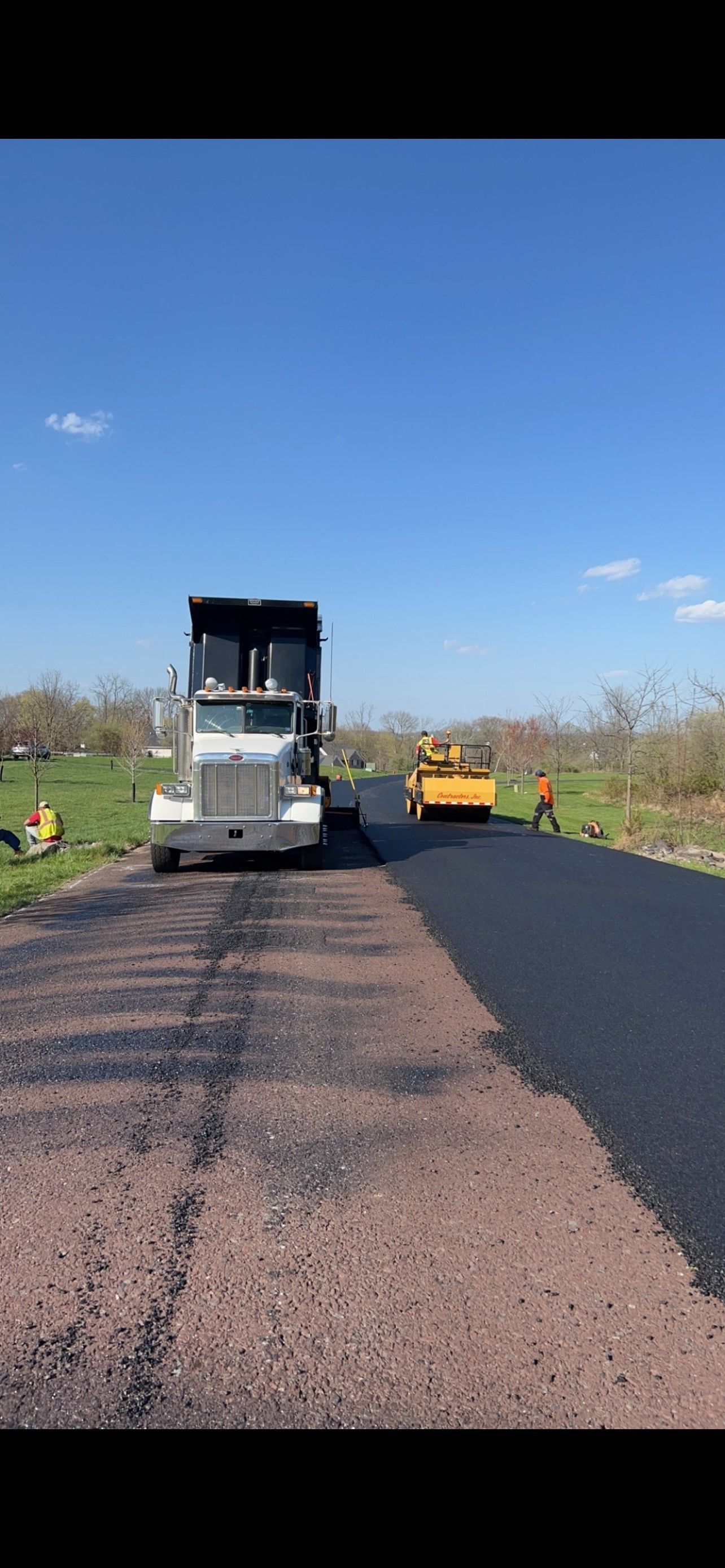 A dump truck and road roller work on a road paving project under a clear blue sky.