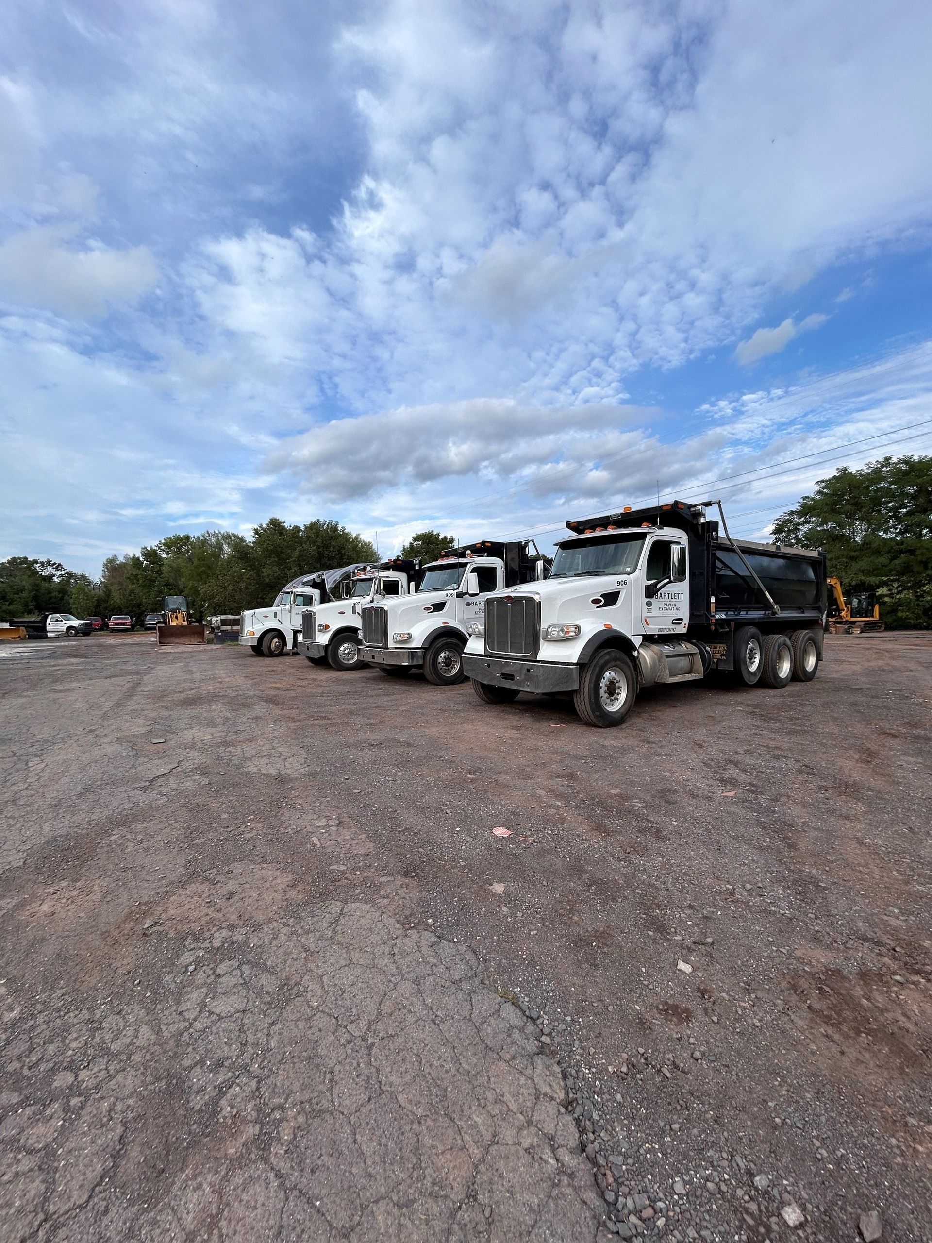 A line of white dump trucks parked on a gravel lot under a cloudy blue sky.