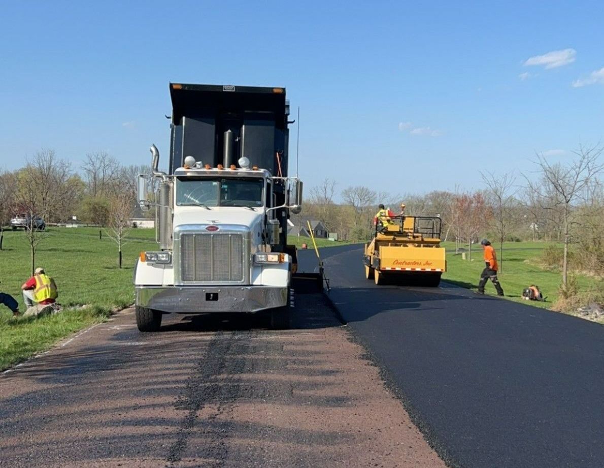 A construction crew paves a rural road with a dump truck and a yellow roller under a clear blue sky.