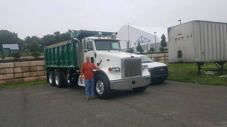 A person standing next to a white dump truck with a green bed in a paved parking lot near a trailer and large building.