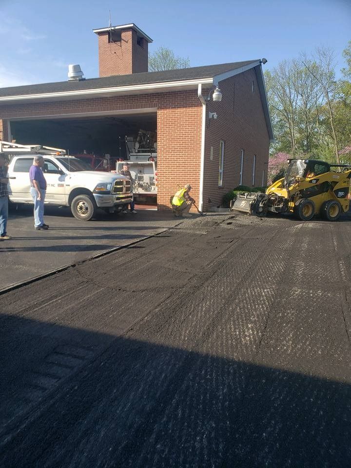 Workers pave a driveway in front of a brick fire station, using a yellow skid-steer loader and a white pickup truck.
