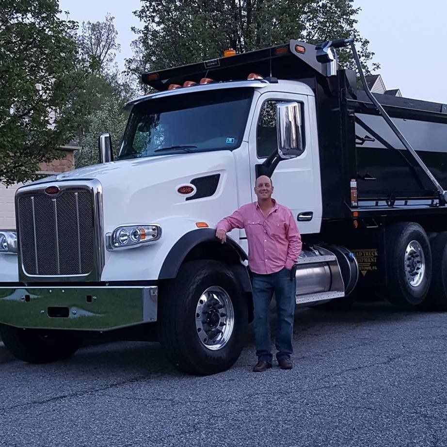 A person wearing a pink shirt and jeans stands smiling next to a white Peterbilt dump truck outdoors on an asphalt road.
