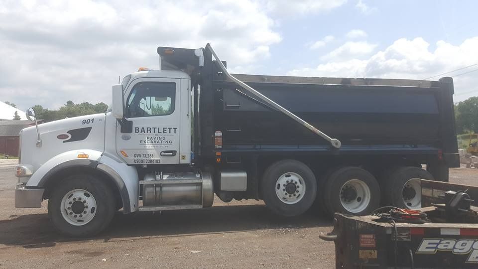 A white dump truck with a black cargo bed parked on a gravel lot under a cloudy sky.