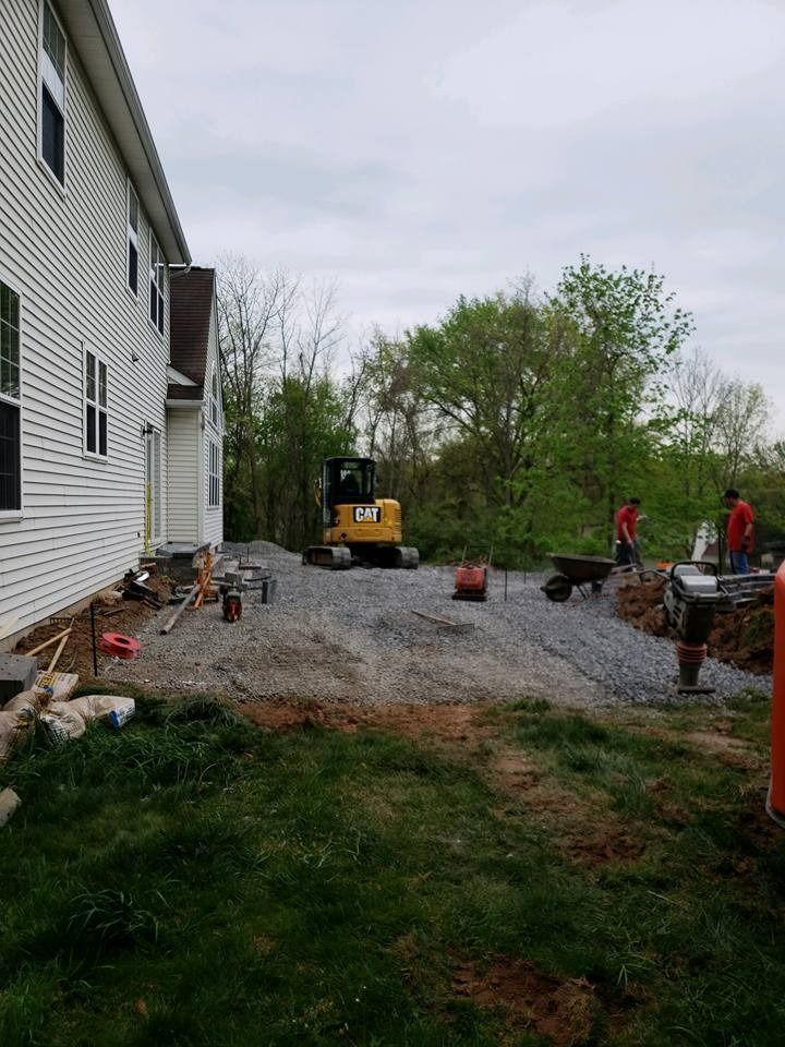 Construction workers with machinery prepare a gravel area next to a white-sided house.