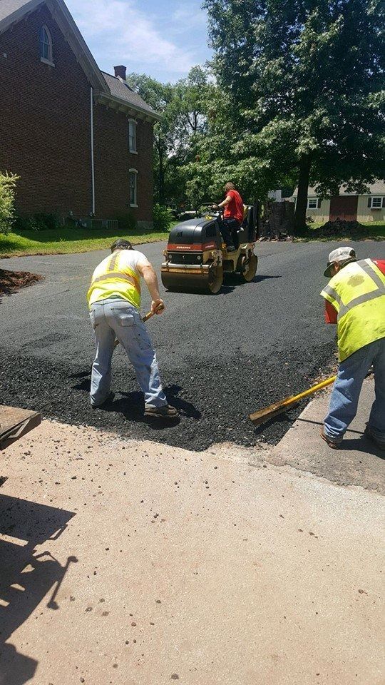 Three workers in high-visibility vests pave a driveway, with one operating a small steamroller near a brick house.