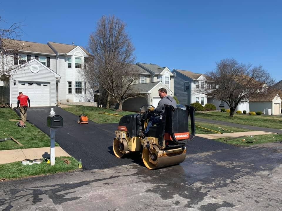 A worker operates a yellow and black asphalt roller to pave a residential driveway in front of a two-story house.