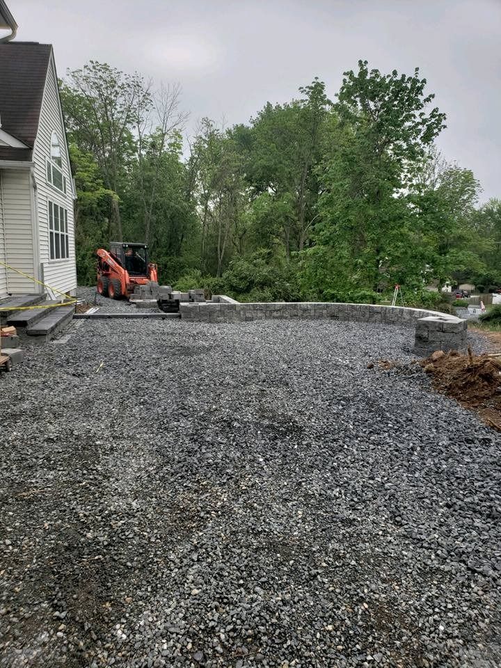An orange skid steer sits in a backyard next to a house with a newly constructed stone retaining wall and gravel base.