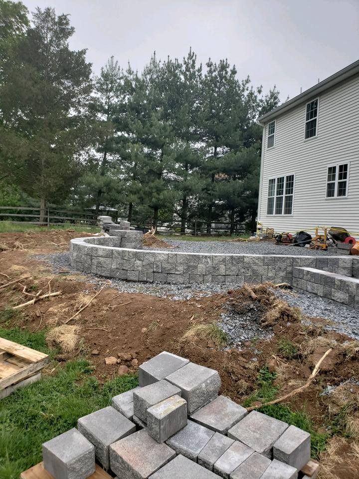 A gray stone retaining wall under construction in a residential backyard next to a light-sided house, with pavers nearby.