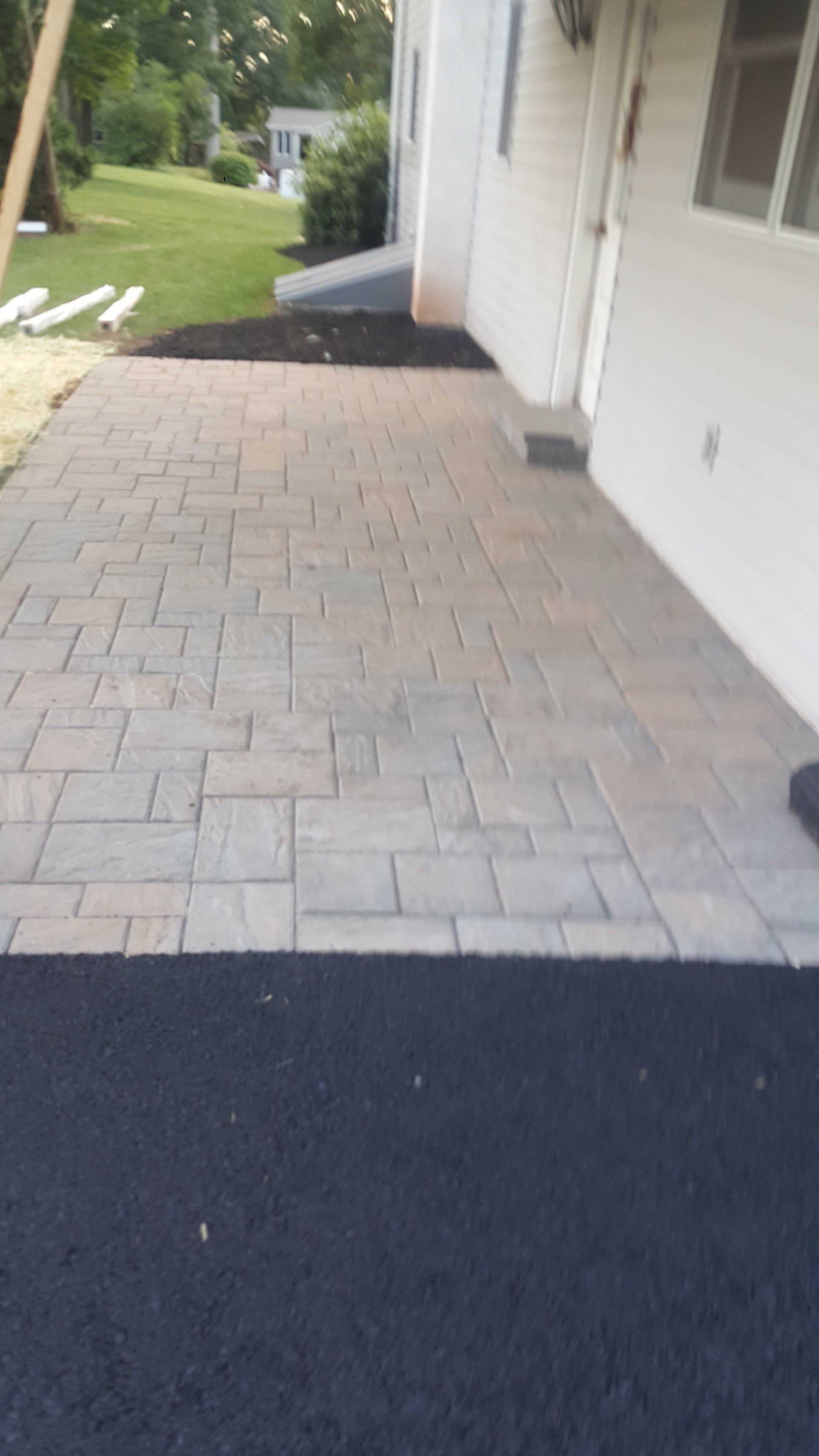 A paved patio made of rectangular stone tiles leads to a white building exterior, with a section of fresh asphalt in front.