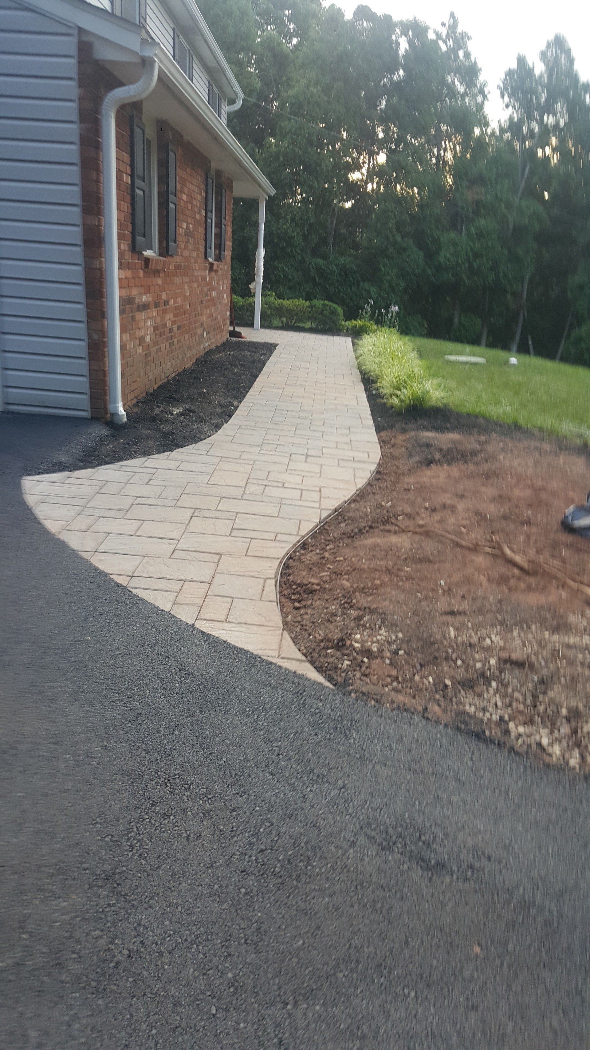 A light-colored brick paver walkway curves along a brick house beside an asphalt driveway and a grassy lawn.