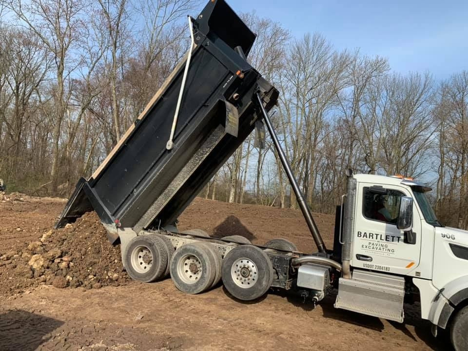 A white dump truck with a raised black bed, unloading a pile of dirt at a construction site with trees in the background.
