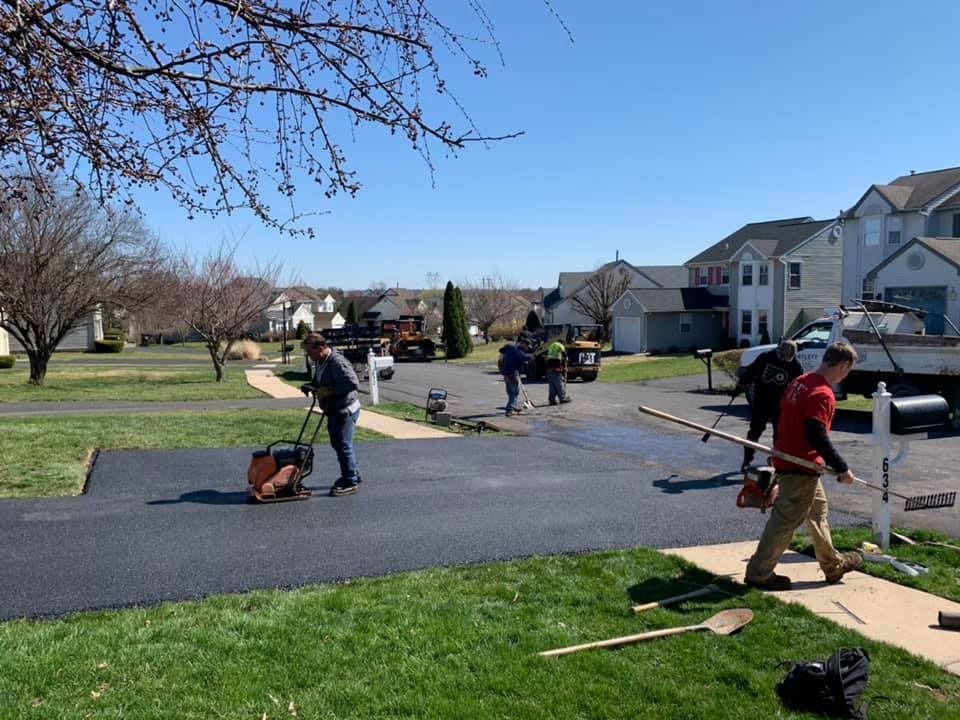 Workers paving a suburban driveway on a sunny day, using machinery and tools to smooth the asphalt.