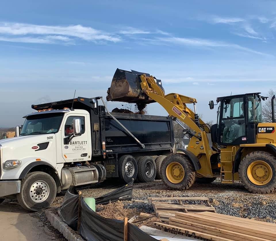 A yellow Caterpillar loader dumps dirt into the black bed of a white dump truck on a sunny construction site.