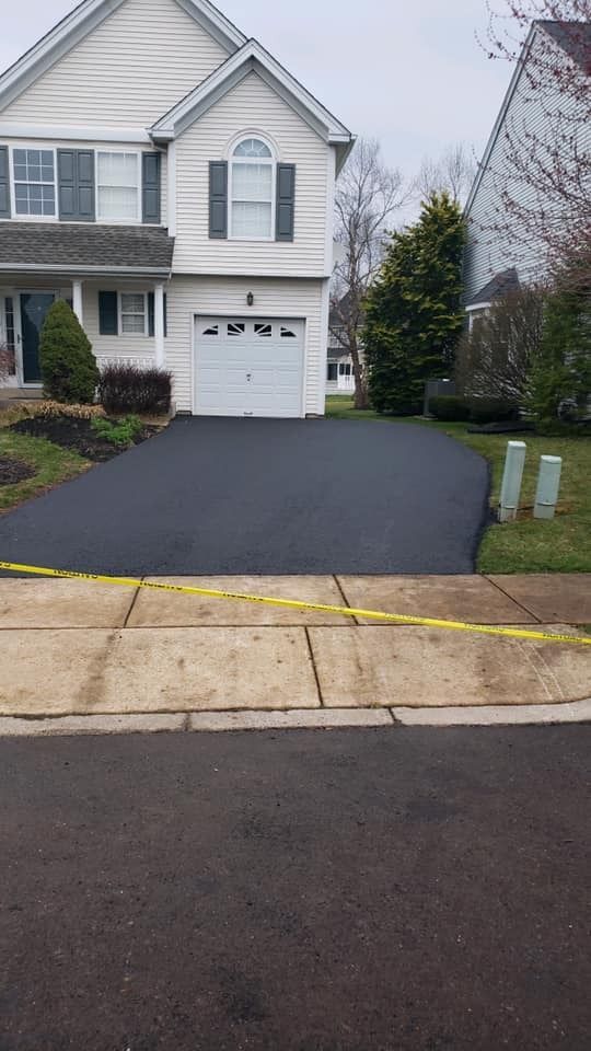 A newly paved asphalt driveway in front of a light-colored suburban home, with yellow caution tape across the sidewalk.
