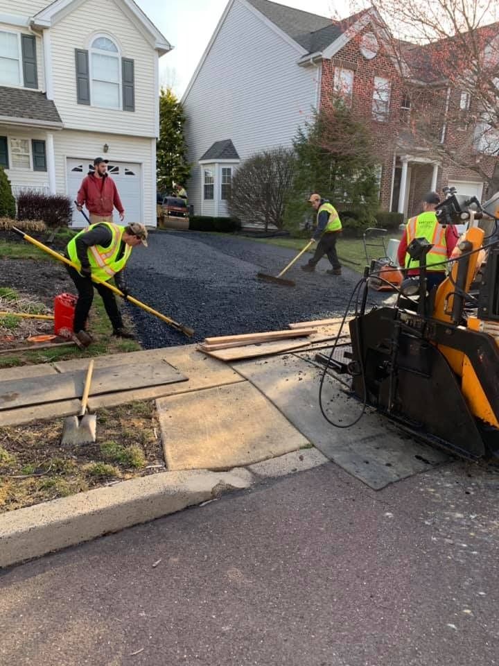 Construction workers in safety vests use rakes to spread fresh black asphalt for a new driveway in a residential area.