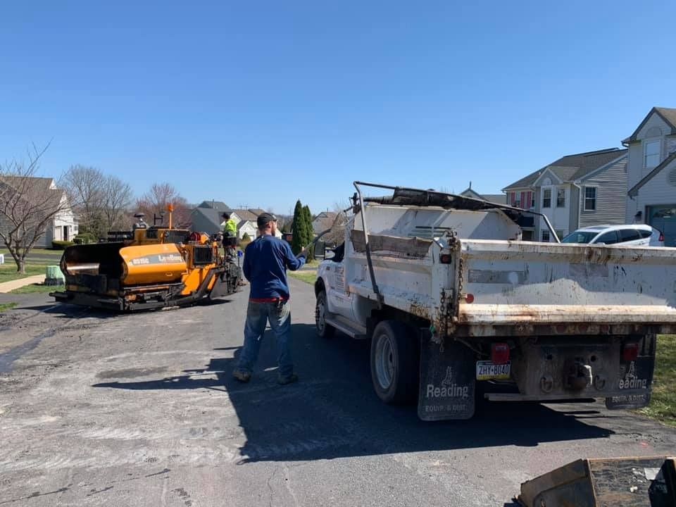 Workers operate a yellow paving machine and dump truck on a residential street during a road construction project.