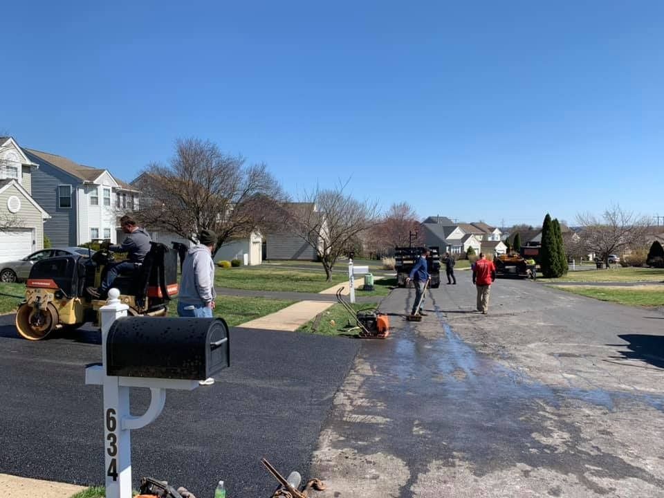 Workers operate machinery to pave a driveway with new asphalt on a sunny day in a residential neighborhood.