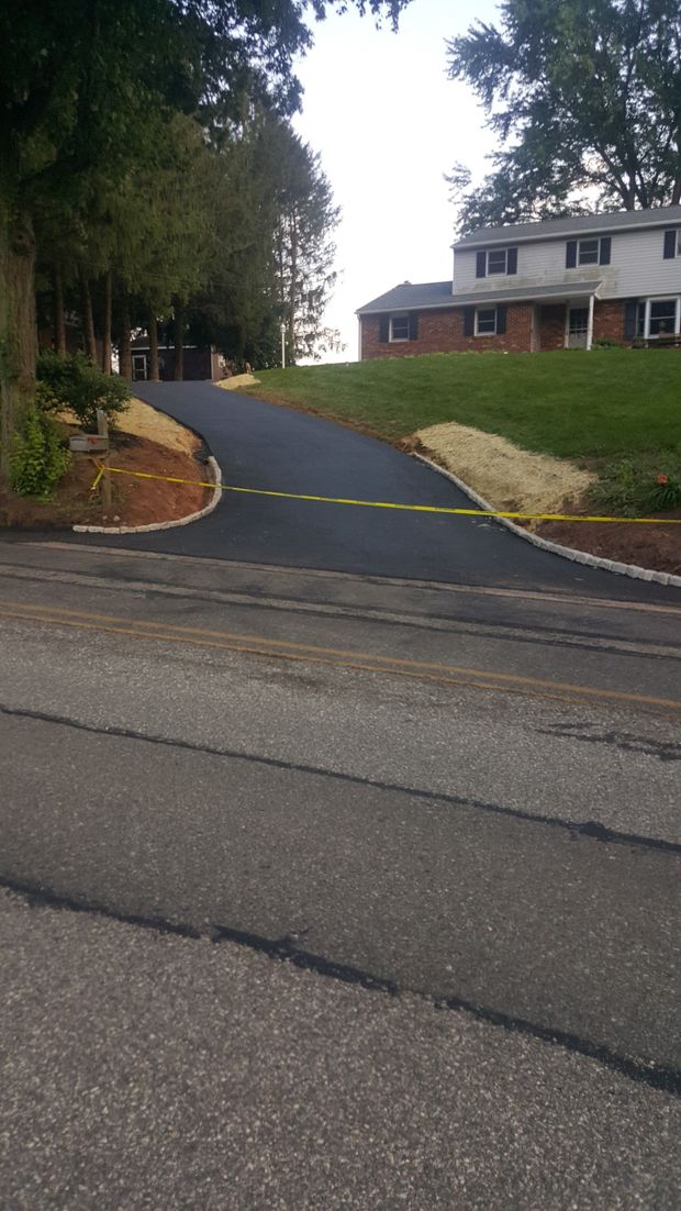 A newly paved asphalt driveway with stone curbing, blocked off by yellow caution tape, leading to a house on a grassy hill.