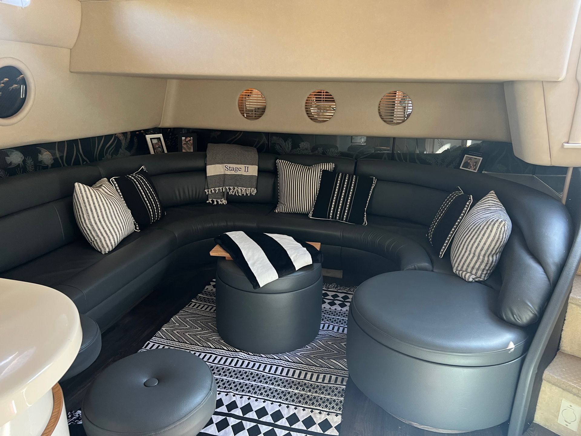 Interior of a boat with a cream colored, curved leather couch and two round ottomans; dark flooring - After