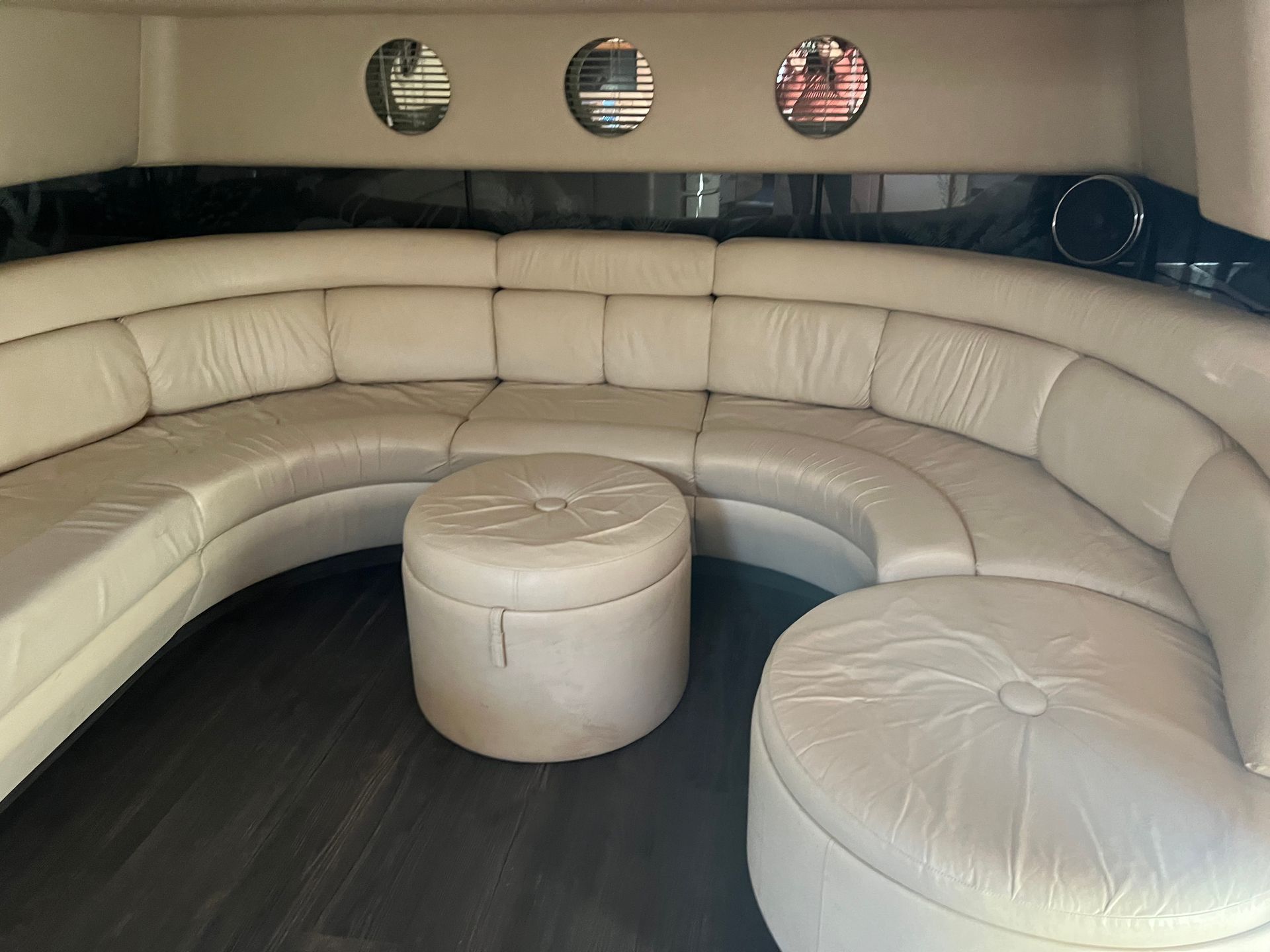 Interior of a boat with a cream colored, curved leather couch and two round ottomans; dark flooring - Before