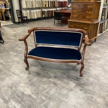 Antique blue velvet loveseat with wood frame, set in a store with wood furniture and fabric swatches.