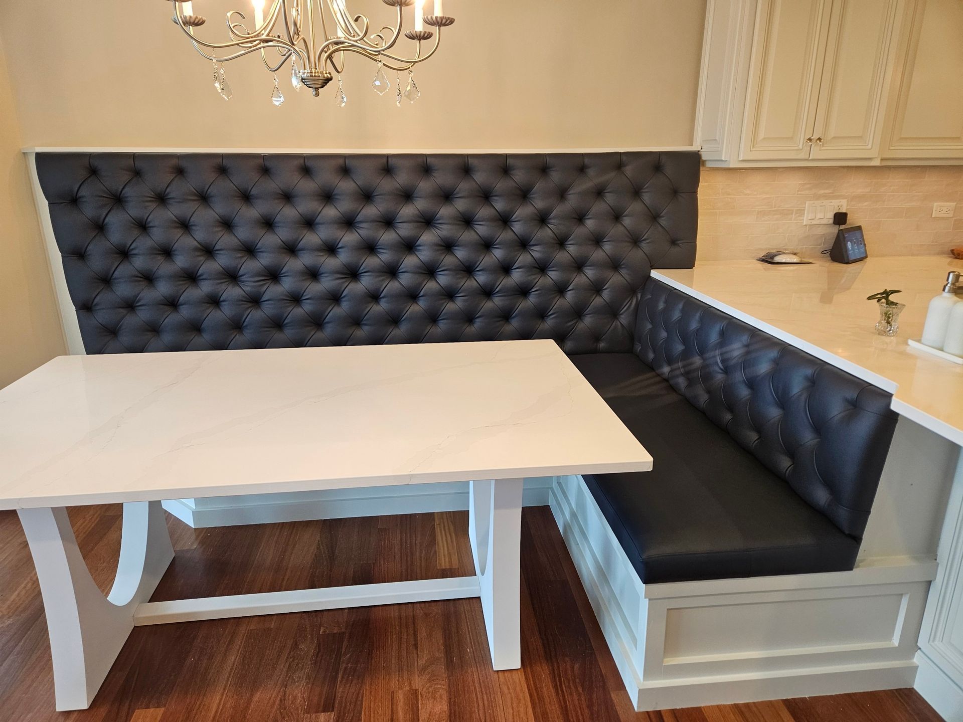 A built-in kitchen corner banquette with a tufted gray back and black seat, white table, and white cabinetry.