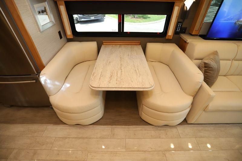 An RV interior featuring a dinette table and two cream-colored bench seats, with a large window and another couch visible.