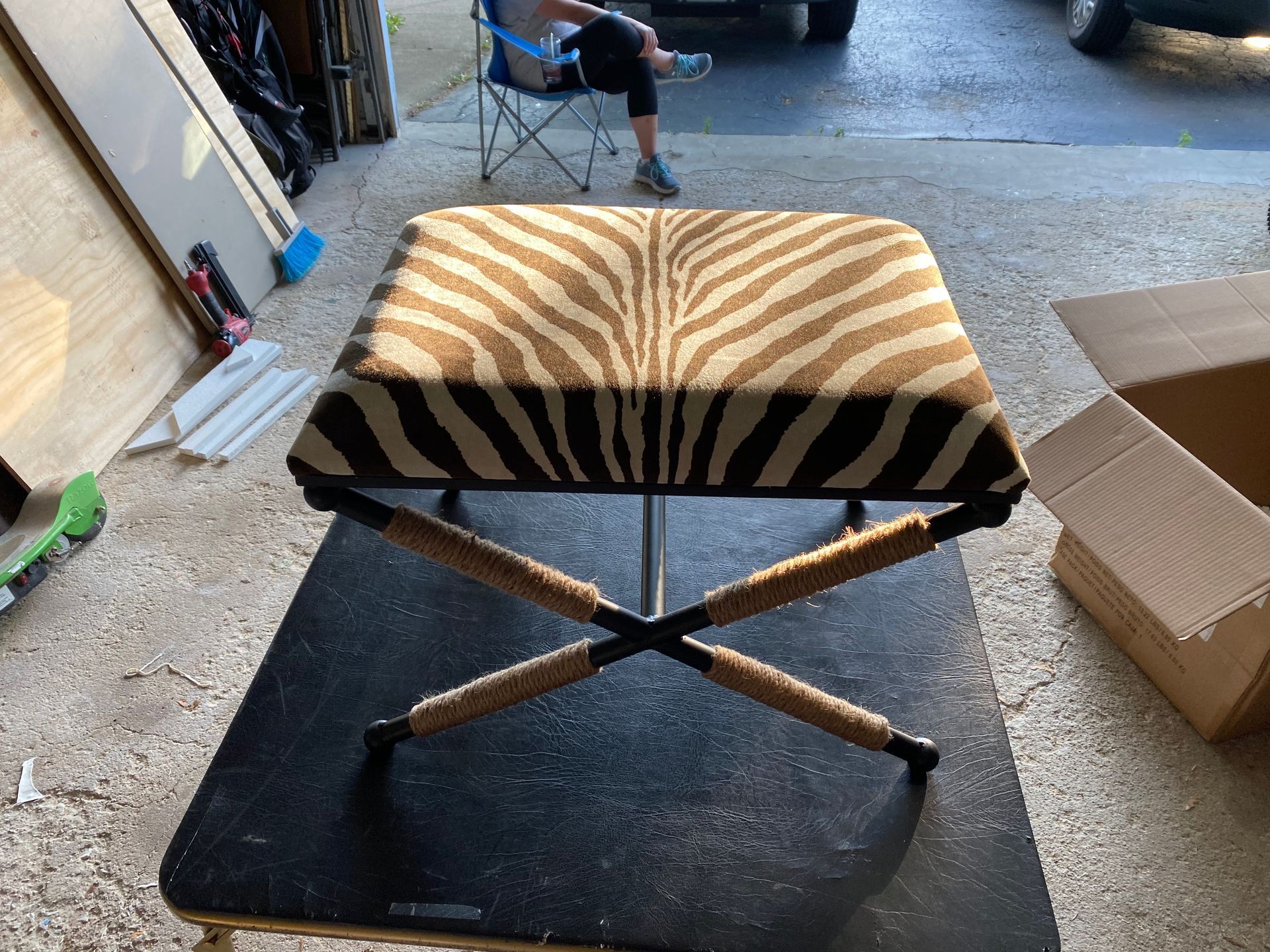 Zebra-print ottoman with black crisscross legs, on a black table outdoors, person in background.