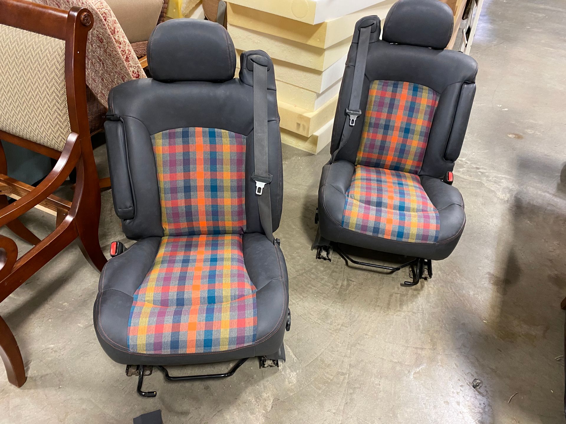 Two used car seats, likely front seats, with gray leather and plaid fabric inserts. They are on a concrete surface with other furniture visible.