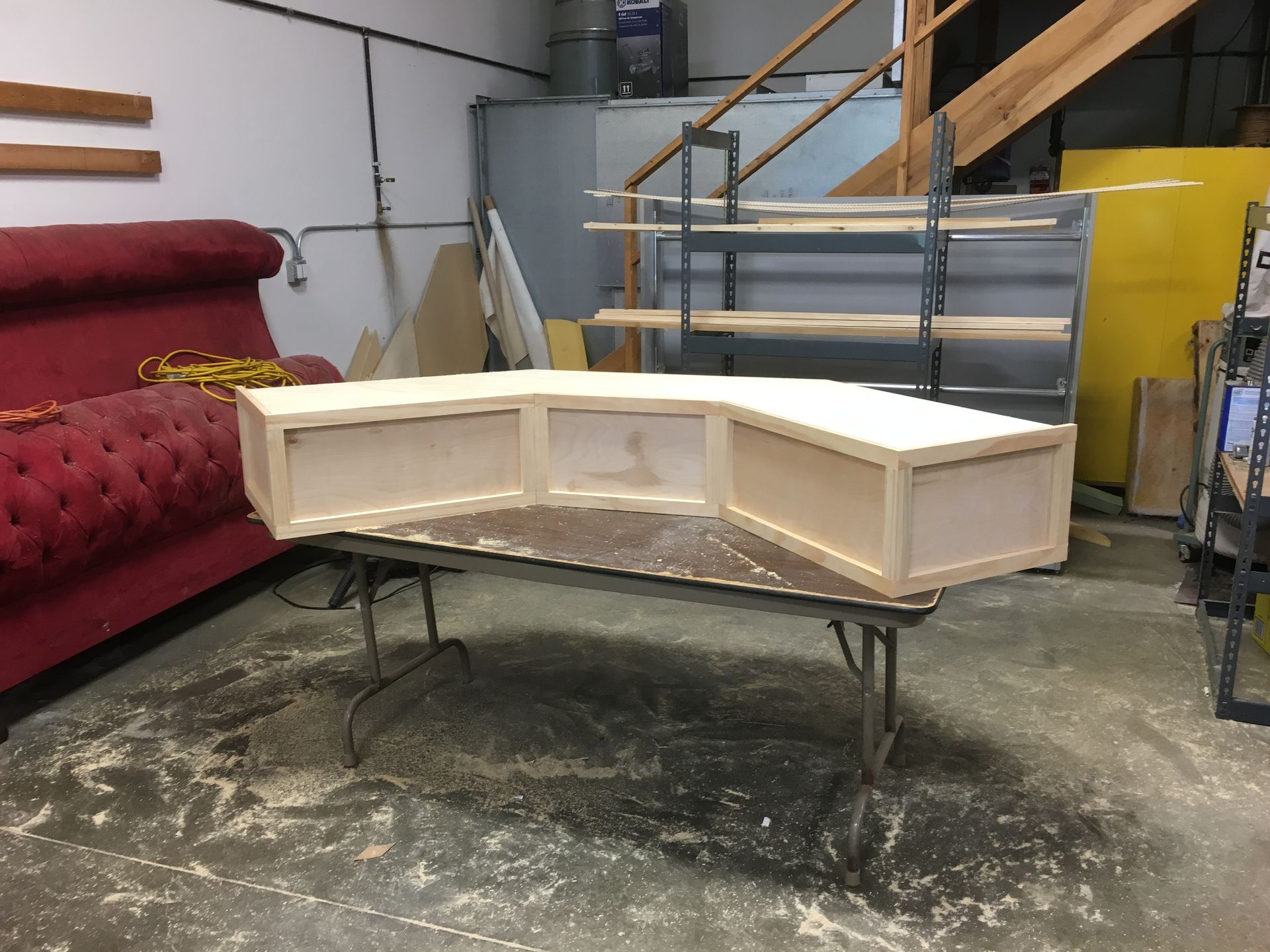 A partially built, light-wood angled cabinet sits on a folding table in a workshop. A red couch and shelves are in the background.