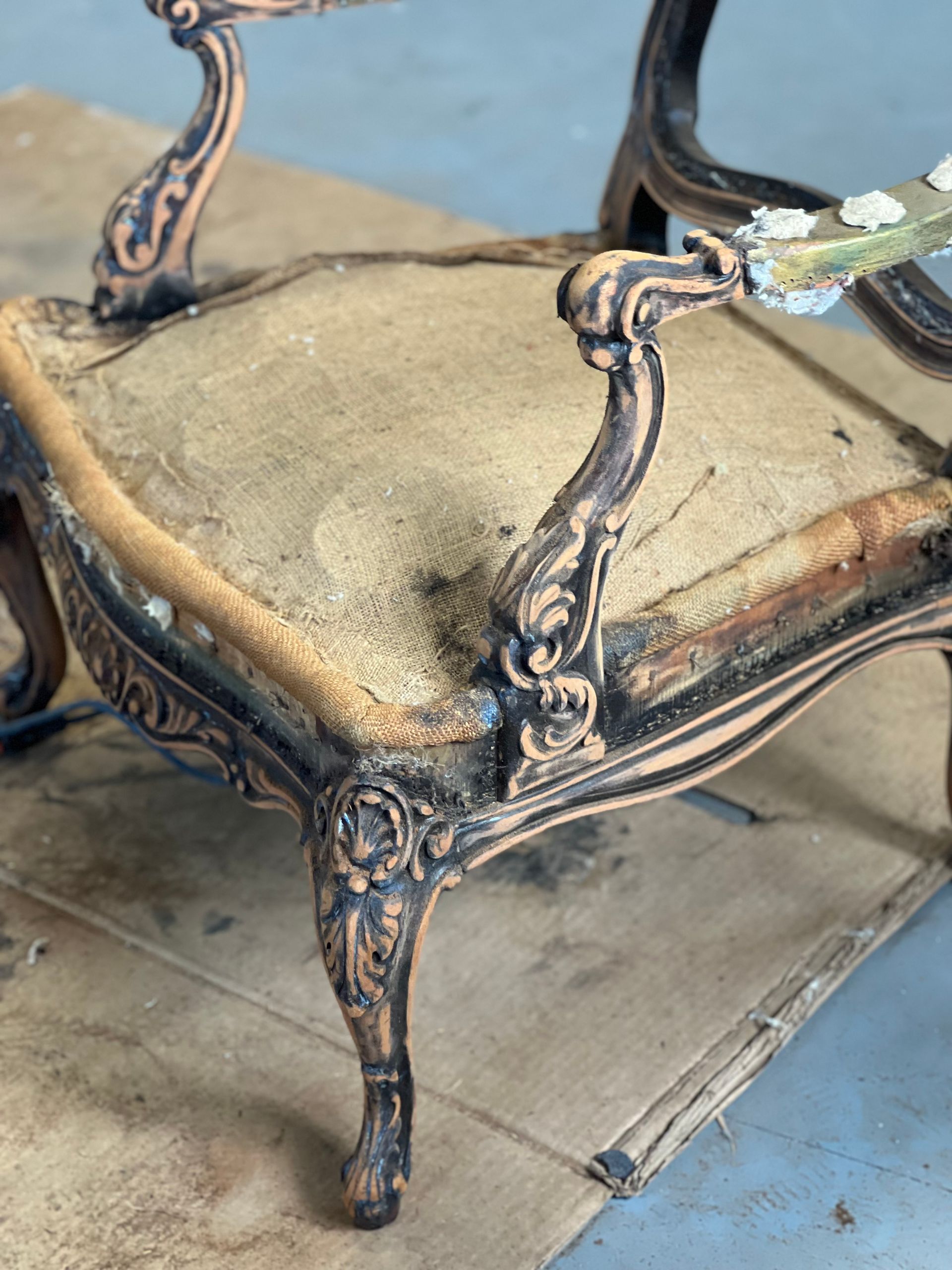 Antique chair with ornate dark brown frame and worn, tan seat. The armrest is curved and detailed.