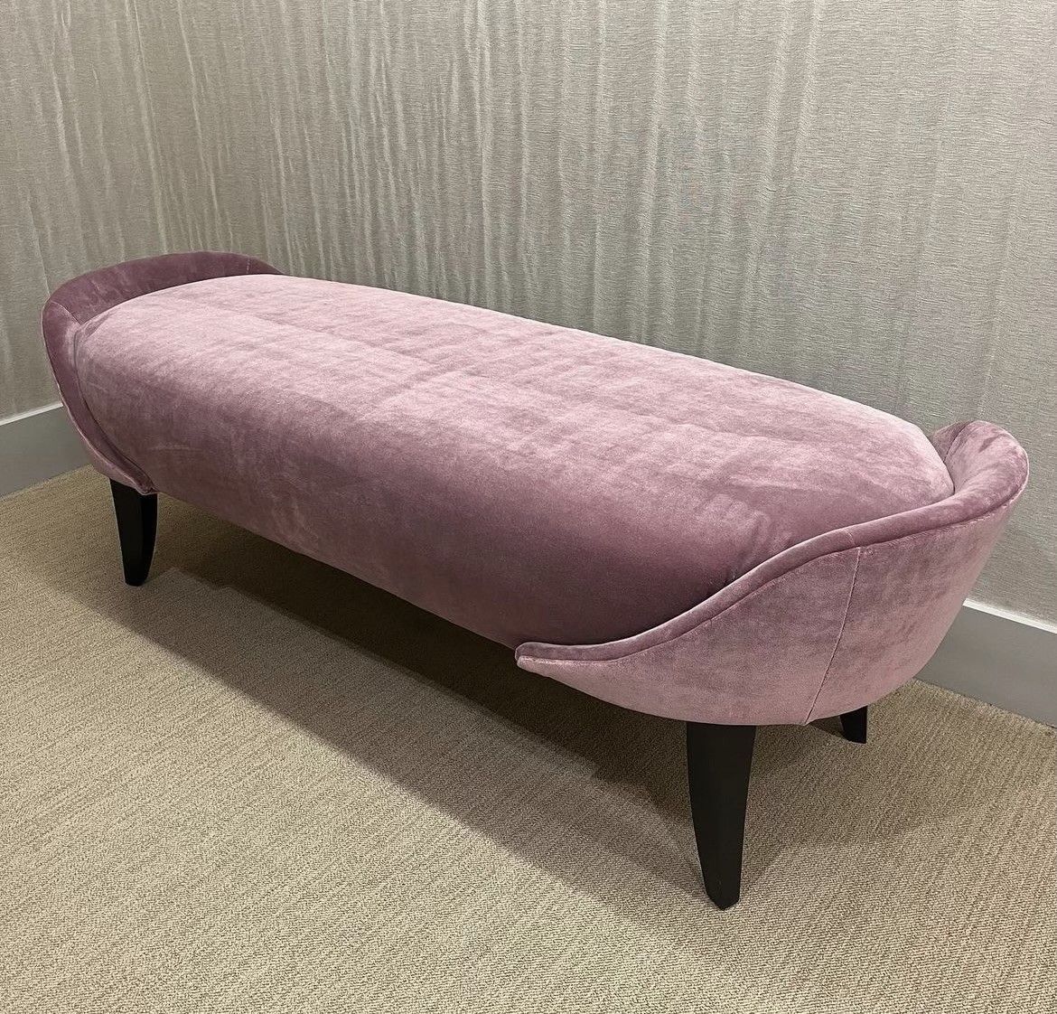 A low, mauve velvet bench with curved ends and dark wooden legs. It sits on beige carpet against a textured wall.