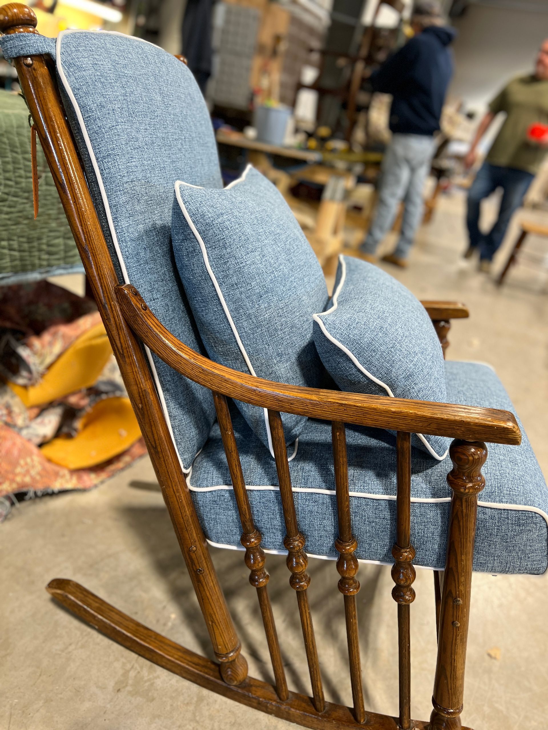Blue upholstered rocking chair with white piping and brown wooden frame, indoors. Two people are in the background.