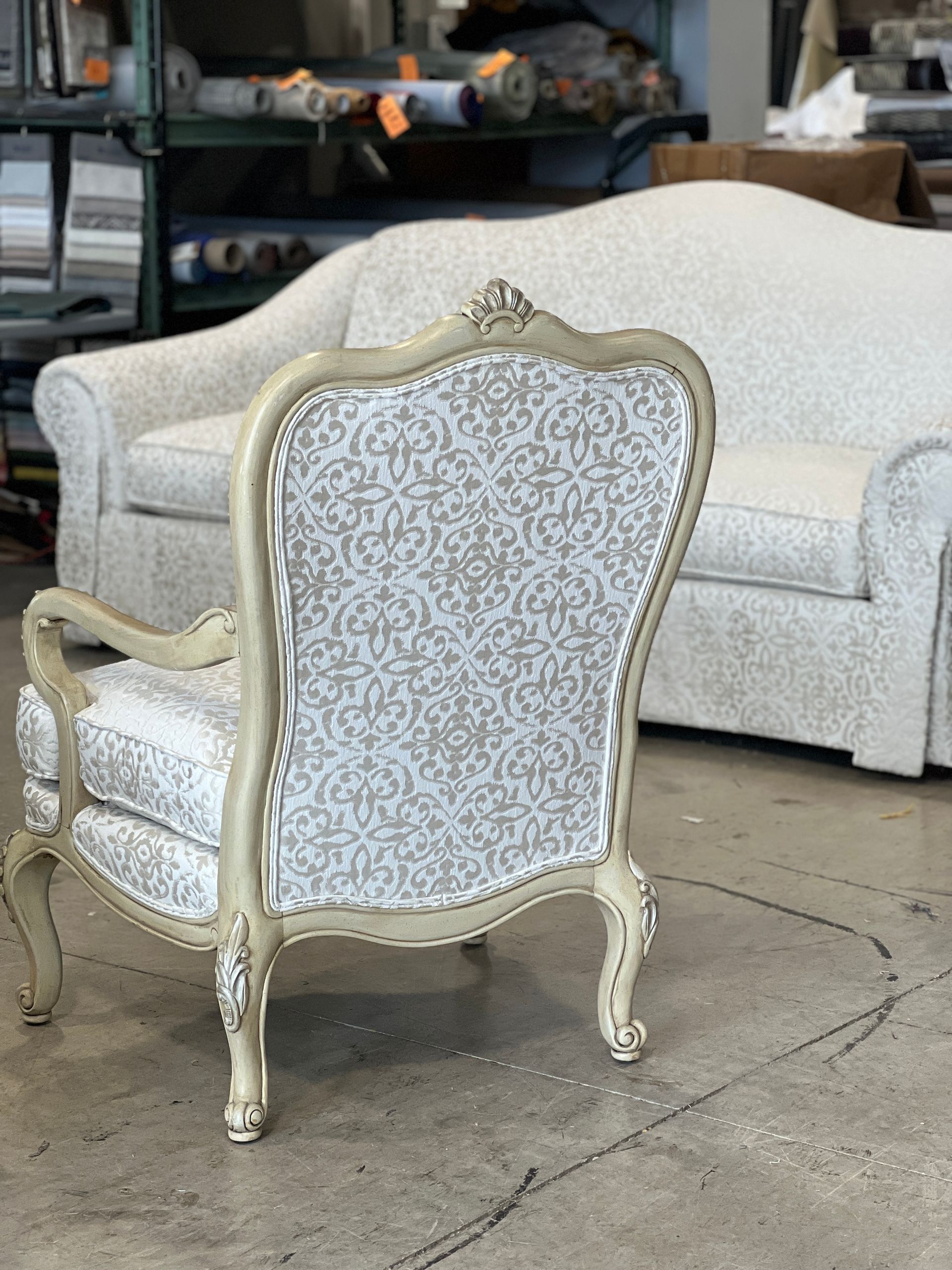 An ornate, cream-framed chair with a patterned back and seat stands in front of a matching sofa. Both are reupholstered in a gray and white fabric.