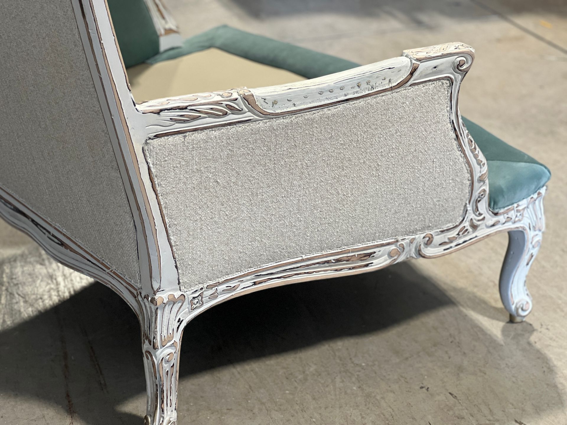 Close-up of an ornate, antique loveseat. It has a light blue cushion, a textured gray back, and a white, distressed frame.