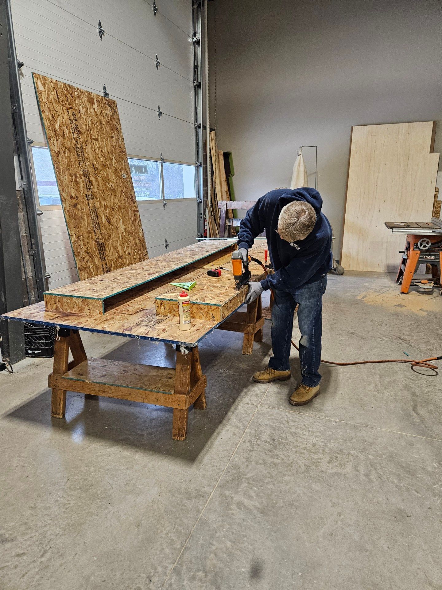 A person in a workshop uses a power tool on OSB boards set on sawhorses. Light shines through a garage door.