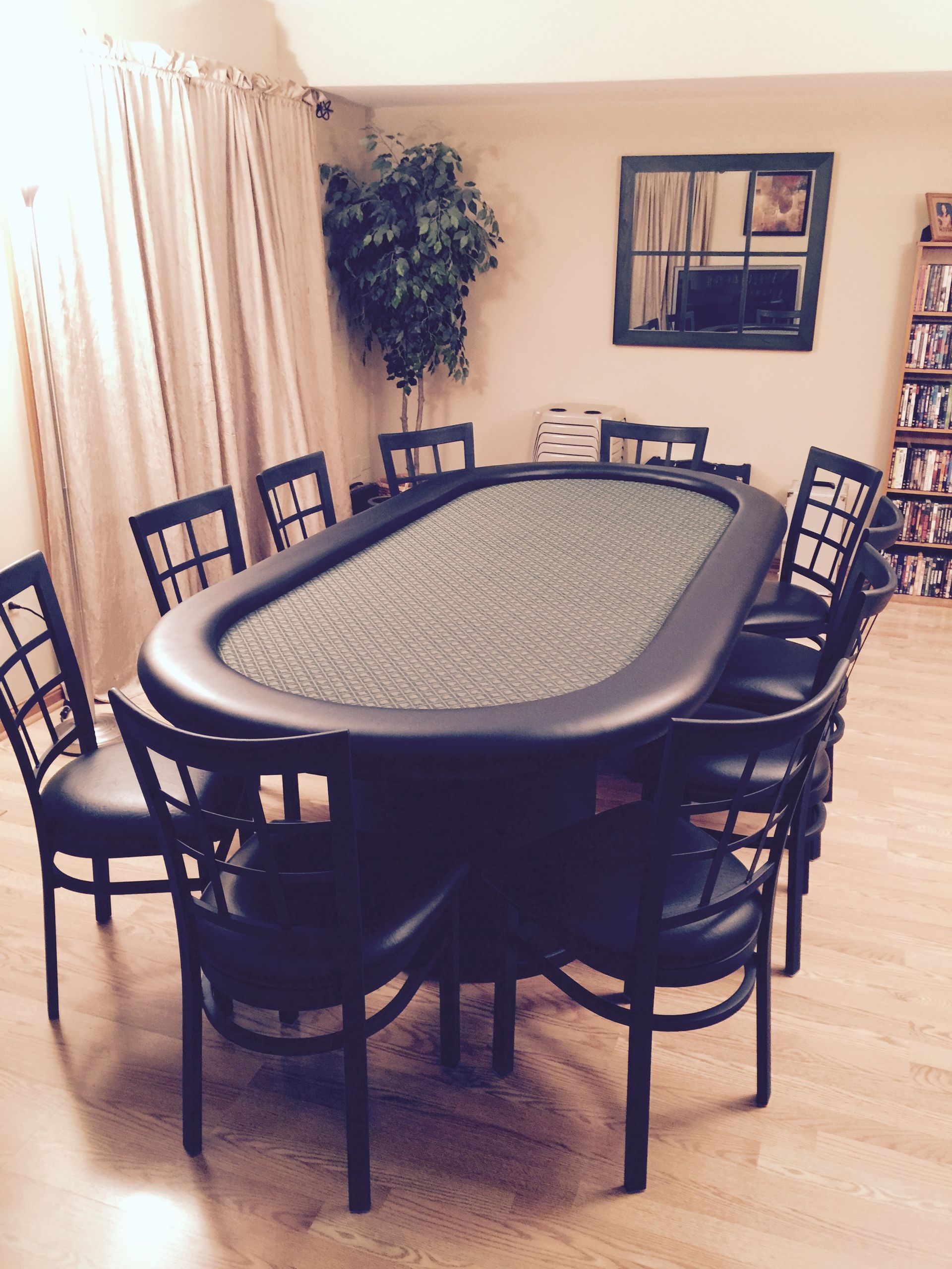 A black poker table surrounded by 11 black chairs in a room with a tree, mirror, and bookshelf.