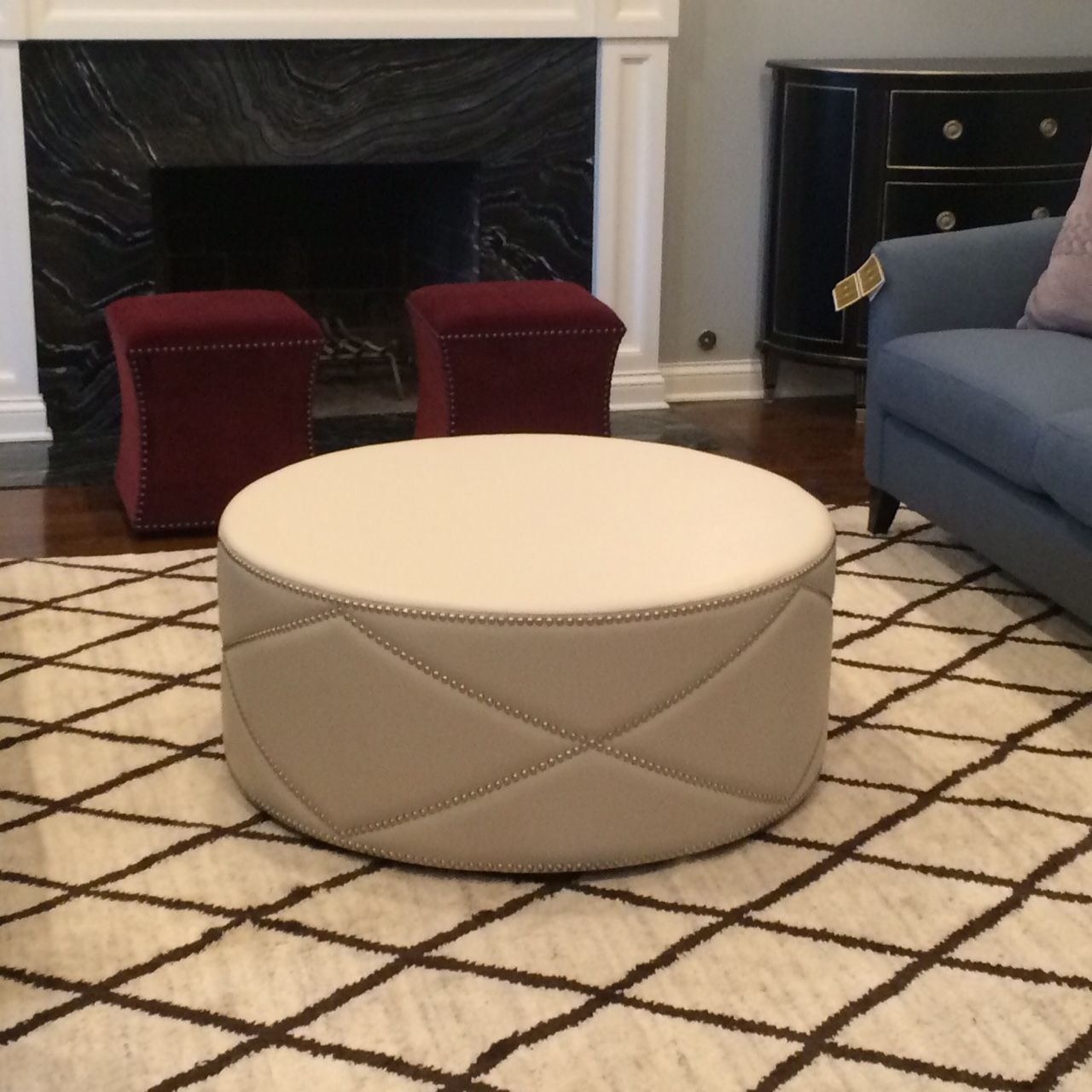 A beige, round ottoman with diamond stitching sits on a patterned rug. Two maroon ottomans and a blue sofa are in the background.