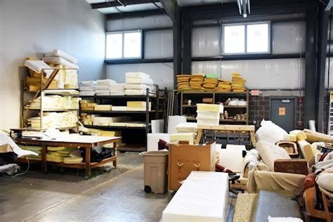 Warehouse interior with shelves of foam, work tables, and various materials, suggesting a furniture or upholstery workshop.
